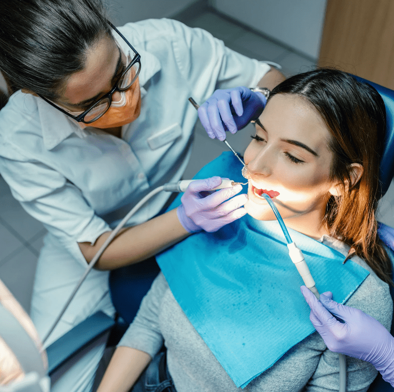 A female patient receiving dental treatment from a dentist wearing a mask and gloves. The dentist uses dental tools while an assistant holds a suction device.